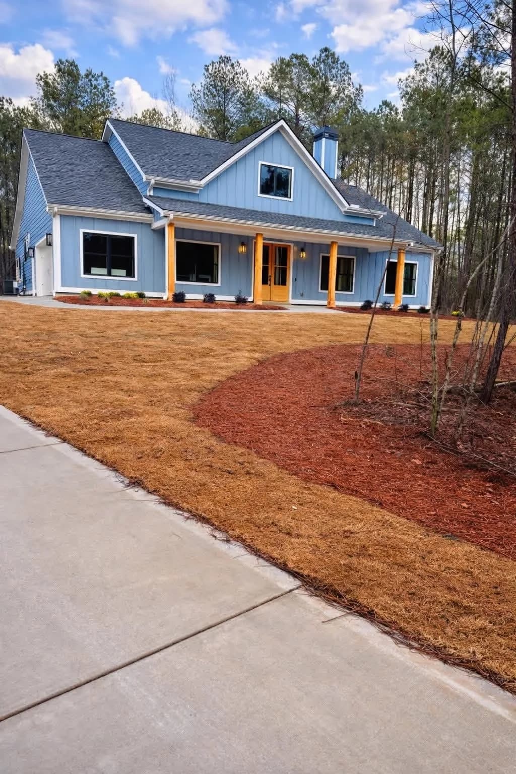 New construction landscaping at a blue farmhouse: fresh grading, mulched beds, and a new concrete drive